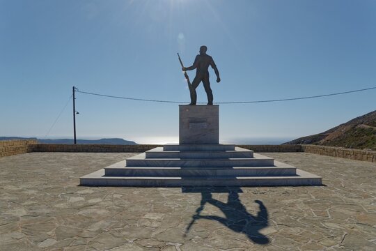 Carpathian statue on a pedestal with outstretched arm and rifle, shadow on the ground, sea in the background, Menetes, Karpathos, Greece