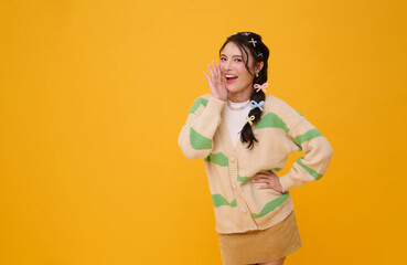 Smiling Asian woman making shouting gesture with hand near mouth in casual fashion outfit expressing communication positivity playful energy and modern lifestyle isolated on yellow background