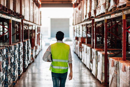 A lone male warehouse worker, seen from behind, walks down an aisle of a warehouse while holding a hard hat. He is wearing a bright safety vest.