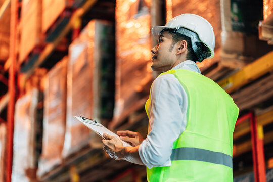 A young male warehouse worker in a white hard hat and bright safety vest looks up at the inventory on the shelves while holding a clipboard. He is focused and attentive to the work.