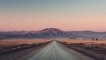 A long, straight desert road vanishes towards a large, imposing mountain range under a soft, pastel sunset sky