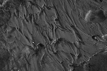 High angle close up view of a textured surface composed of sand and mud. The grayscale image reveals a pattern of ridges, grooves, and swirls, suggesting the effects of water or wind erosion.