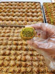 A hand wearing a transparent glove holds a piece of baklava topped with finely ground green pistachios (Pistacia vera). In the background, a large tray displays multiple pieces of neatly arranged bakl