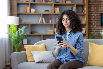 Young woman sitting comfortably on a gray sofa at home, engrossed in checking social media or...