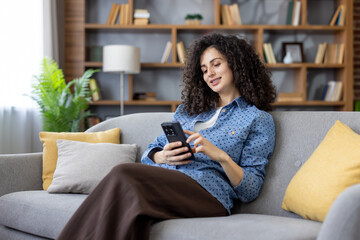 Obraz premium Young woman with curly hair sitting comfortably on a gray sofa in her living room, smiling and scrolling through content on her mobile phone