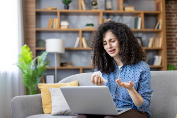 Upset young woman experiencing emotional distress, holding a tissue, and crying while using a laptop for communication at home, conveying concepts of sadness, bad news, despair, and online support