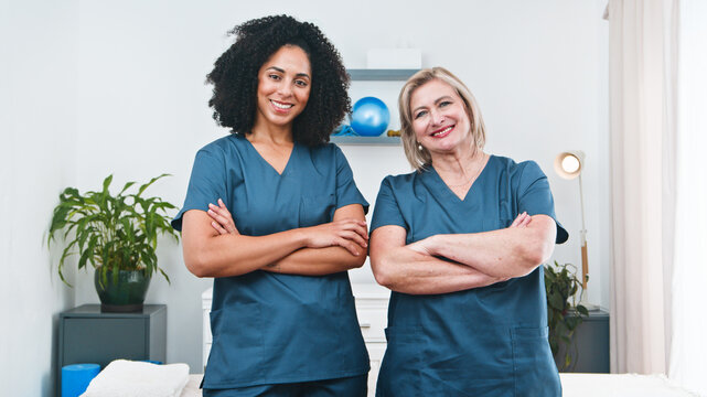 Two Female Physiotherapists Stand Confidently With Crossed Arms In A Clinical Therapy Setting