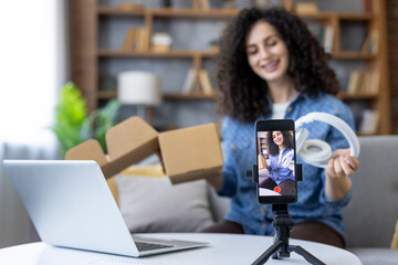 Young woman vlogger unboxing new headphones at home, filming a live product review on her smartphone for social media, smiling as she presents the gadget to viewers