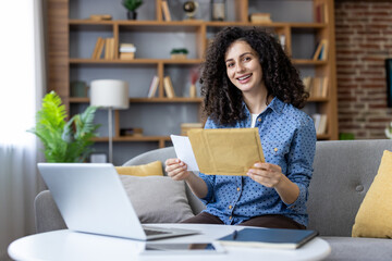 Young woman with curly hair smiling as she opens an envelope on a sofa beside her laptop in a cozy home office, receiving happy personal or work-related mail