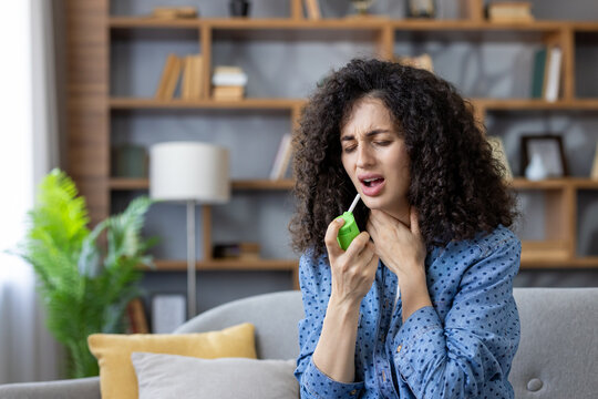 Young woman on a couch spraying throat medicine into her mouth, wincing in discomfort as she self-treats sore throat and cold symptoms at home