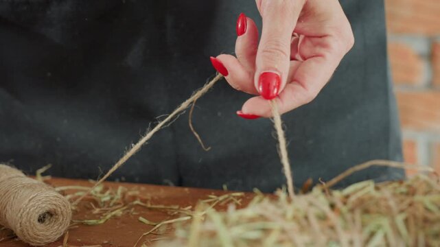 Close up of female hands with red nail polish tying twine around straw wreath on wooden table, focusing on handmade crafting, rustic decoration, and artisan detail in floral arrangement preparation