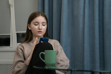 Sitting comfortably by the window with a coffee mug on a glass table nearby, a young girl is using her smartphone, browsing social media or texting
