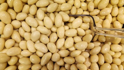 Pale yellow, almond-shaped chocolates are clustered together. They have a glossy surface, indicating a smooth, possibly sugar-coated finish. A metal scooping tool is partially visible, suggesting acce