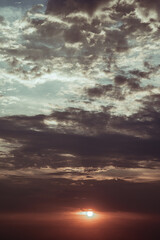 A vibrant sunset is captured as the sun dips below a dramatic sky filled with dark and light grey clouds. The clouds showcase a mix of textures and tones, creating depth and visual interest
