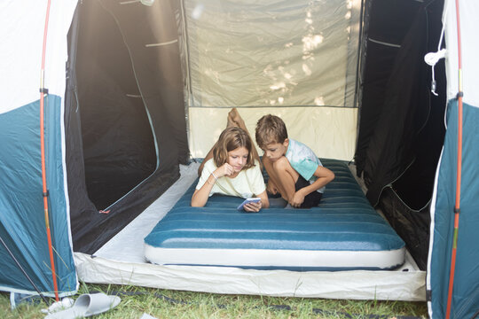 Camping trip with family: children enjoying time inside a tent, bonding over a smartphone in the great outdoors - Powered by Adobe