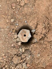 A top-down shot of a concrete post or pillar partially buried in the ground, surrounded by red-brown soil and small rocks. The post has a hexagonal shape with a hole in the center.