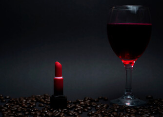 Red lipstick in black case standing among coffee beans beside glass of red wine on dark background. Elegant lifestyle concept with beauty and luxury mood.