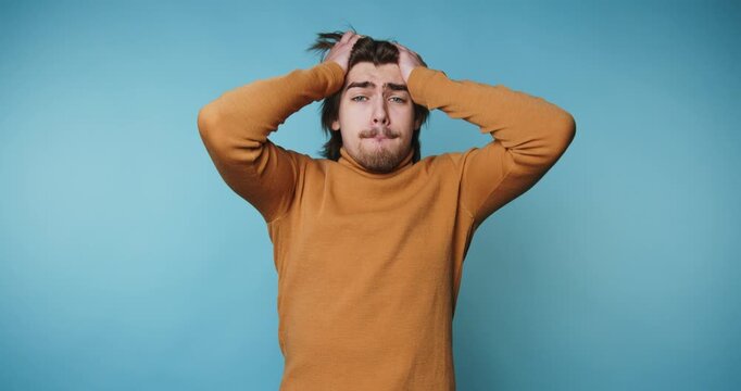 footage of young man expresses frustration by holding his head, capturing a relatable moment of stress on blue background