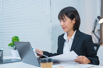  A beautiful businesswoman in a suit is looking at a computer screen while talking on her mobile phone. She is sitting in an office and using modern technology.