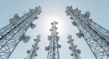 Low angle view of multiple cell towers against a bright sky with telecommunication equipment present