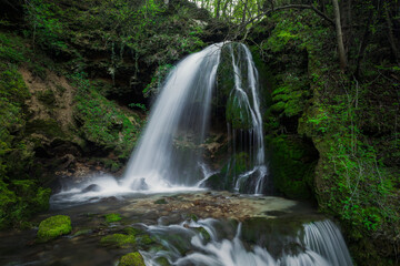 Fototapeta premium A beautiful waterfall cascades down rocky terrain, surrounded by vibrant green plants and trees in a peaceful setting.