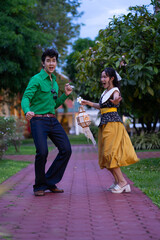 Joyful couple dances on brick path in park, with man in green shirt and jeans, and woman in yellow dress holding lantern