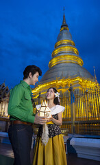 Couple in traditional attire holds lantern in front of illuminated temple at dusk, creating serene and cultural atmosphere