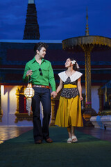 Couple joyfully walks hand in hand at temple during evening, holding lantern. woman wears yellow skirt and man green shirt