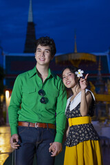 Smiling couple stands in front of temple at night, with woman holding flower. man wears green shirt, and woman wears yellow skirt