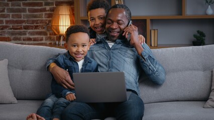 Happy African American family father multitasking two sons cuddling dad surprise dad working with laptop talking mobile phone call at home distant job little boys kids children hugging daddy fun love