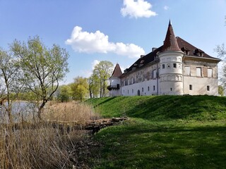 Siesikai Castle in Lithuania is seen on a bright day, surrounded by lush greenery and water. The castle features a white facade with a conical roof tower, red roof tiles, and several windows. 