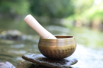 Tibetan singing bowl with mallet on stone near river, closeup