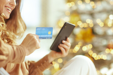 A smiling woman holds a credit card and smartphone, embodying festive holiday online shopping. A...