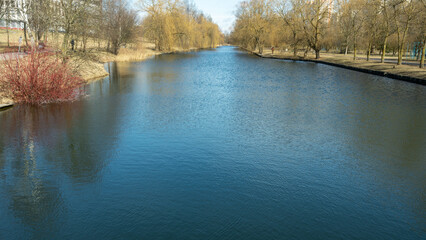 Serene river with leafless trees and walking paths in early spring or autumn urban park. Peaceful canal surrounded by bare trees and grassy banks in tranquil city park with soft ripples on water.