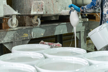 Manual paint pouring process in industrial workshop. White pigment flowing from industrial spout into containers during manual paint production in factory.