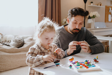 Naklejka premium A 3-year-old girl and her father play with modeling clay in a sunny living room. Unfiltered, authentic photography capturing real family moments of creativity and bonding
