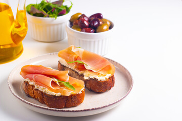 Two bruschettas with prosciutto, ricotta cheese and rucola on ciabatta bread. Olive and spices  decorated.