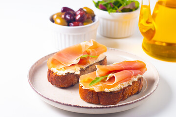 Two bruschettas with prosciutto, ricotta cheese and rucola on ciabatta bread. Olive and spices  decorated.