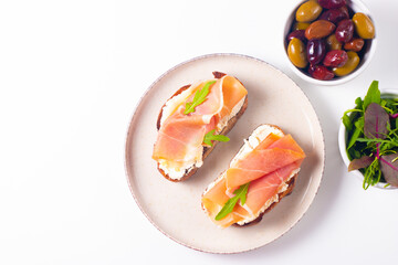 Two bruschettas with prosciutto, ricotta cheese and rucola on ciabatta bread. Olive and spices  decorated.