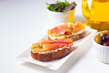 Two bruschettas with prosciutto, ricotta cheese and rucola on ciabatta bread. Olive and spices  decorated.