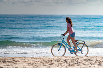 Fototapeta premium A woman is riding a bicycle on a beach. The ocean is in the background. The woman is wearing a pink shirt and shorts