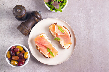 Two bruschettas with prosciutto, ricotta cheese and rucola on ciabatta bread. Olive and spices  decorated.