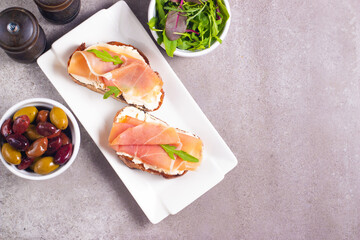 Two bruschettas with prosciutto, ricotta cheese and rucola on ciabatta bread. Olive and spices  decorated.