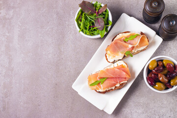 Two bruschettas with prosciutto, ricotta cheese and rucola on ciabatta bread. Olive and spices  decorated.