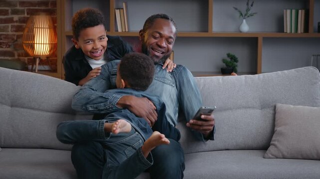 Happy African American family daddy using phone sons jumping running surprise dad cuddling laughing together with mobile smartphone two boys kids children at home enjoying relationship father's day