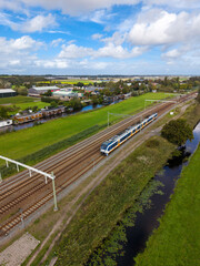 A modern blue white yellow passenger train glides along rural tracks beside a grassy canal, with houses, docks, fields, and trees under a partly cloudy countryside sky.