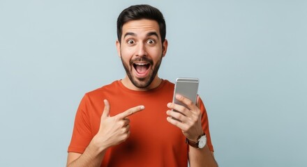 Man with open mouth pointing at cell phone in his hand on a plain blue background excited and happy