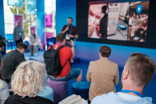 Vibrant conference scene shows presenter on stage, large screen displays, attentive crowd seated on colorful stools, modern event space, blue lighting and energetic atmosphere inviting networking 