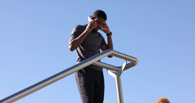 African American man adjusting headphones, stretching quads on bar outdoors, preparing workout
