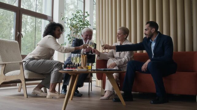 Group of ethnically diverse young and senior men and women sitting at small buffet table, clinking glasses with sparkling wine, celebrating successful presentation
